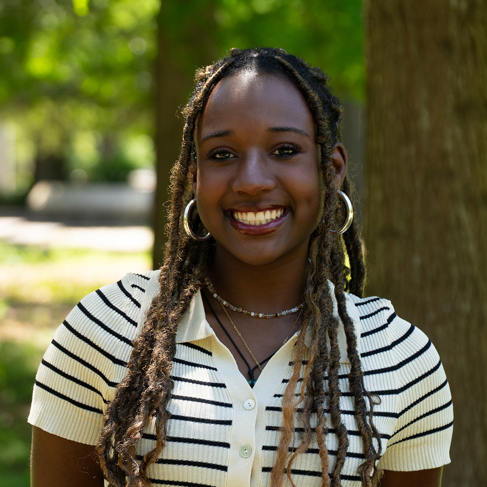 Smiling young woman with braided hair wearing a striped shirt and hoop earrings outdoors.
