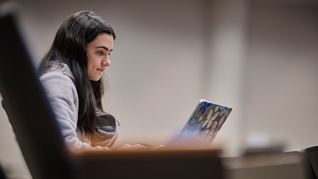 Young woman with dark hair focused on a laptop covered in colorful stickers in a blurred indoor setting.
