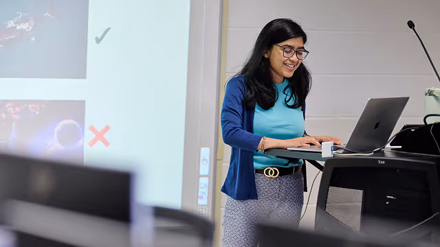 Smiling woman in glasses presents using a laptop beside a screen showing check and cross marks.