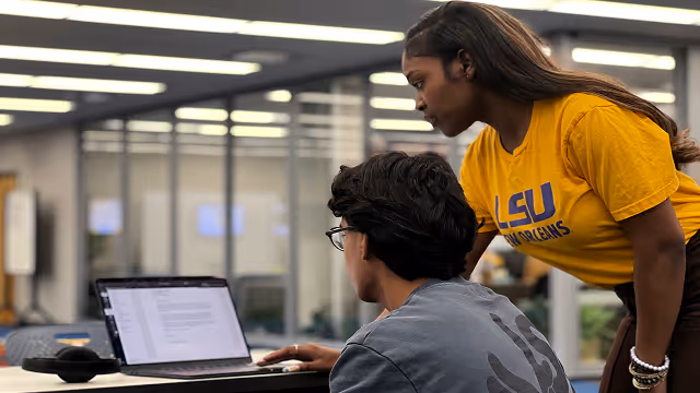 Two students, one standing and one sitting, looking at a laptop screen in a library or study area.
