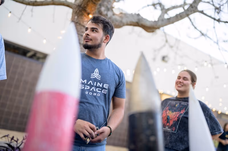Two students stand outdoors near large model rockets, one wearing a Maine Space 2030 shirt.
