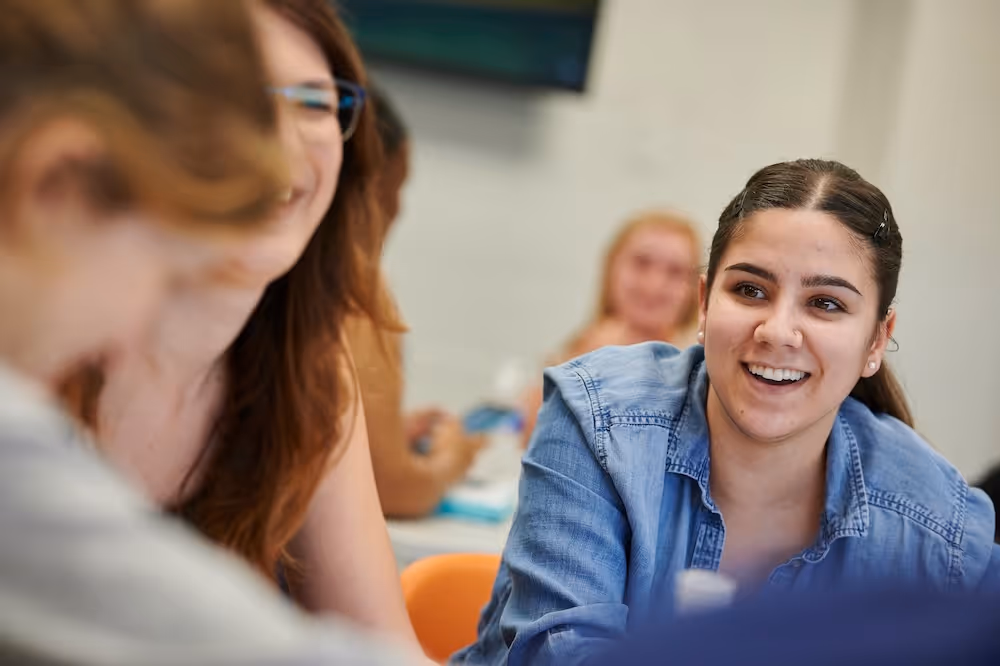 Smiling young woman in denim shirt talking with peers in a casual indoor setting.