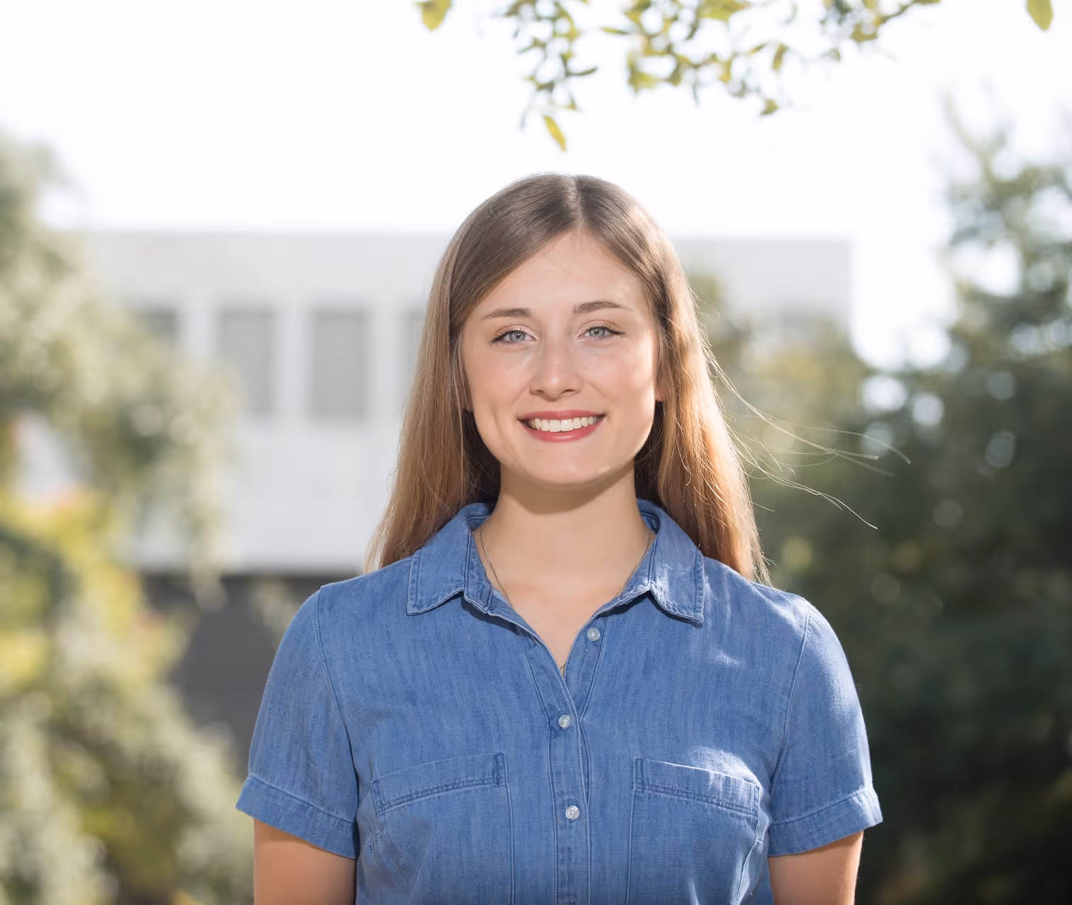 Young woman with long brown hair wearing a blue denim shirt smiling outdoors with trees and a building behind her.