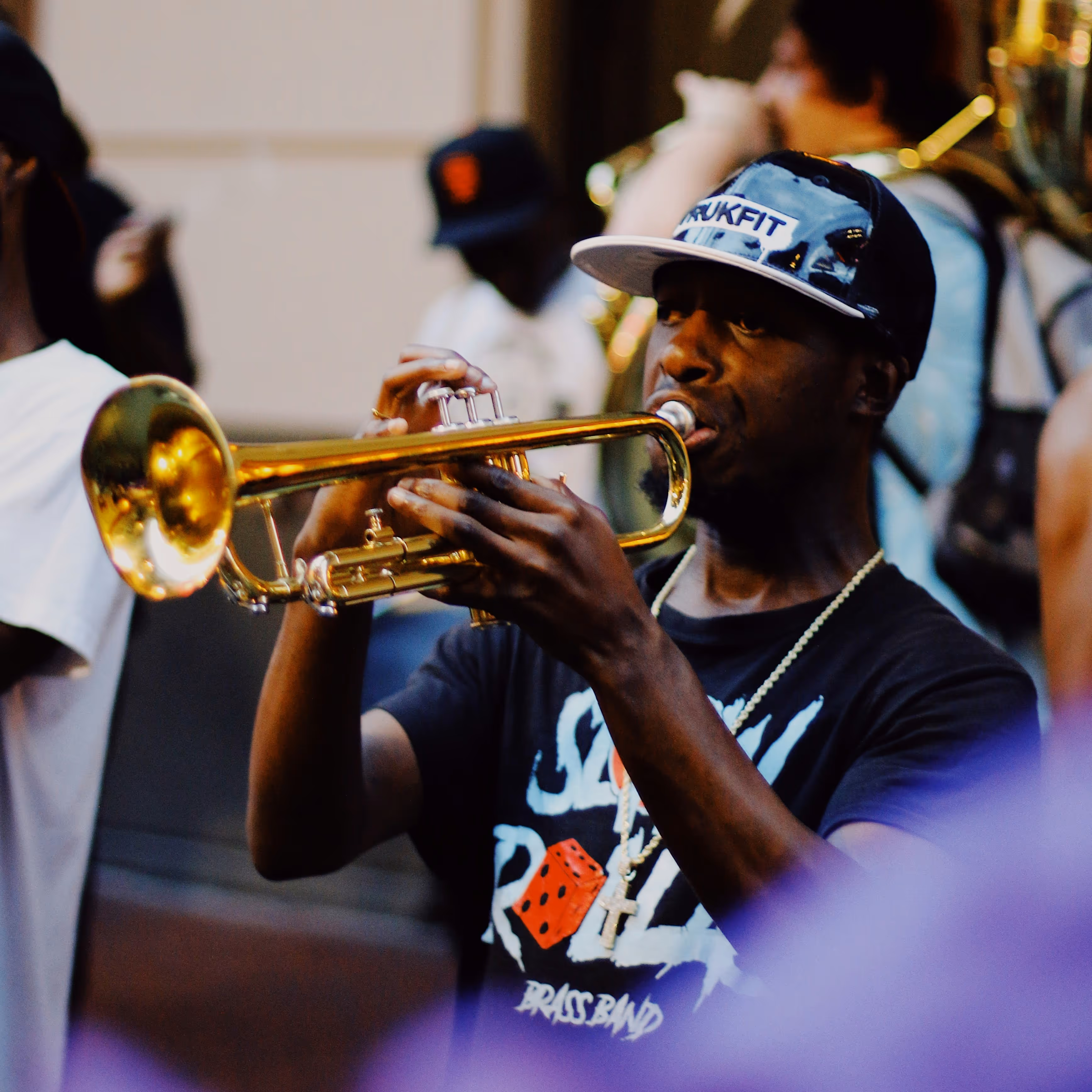 Man wearing a cap plays a golden trumpet during an outdoor musical performance.