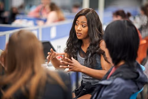 Woman with long hair explaining something to two people in a busy indoor setting.