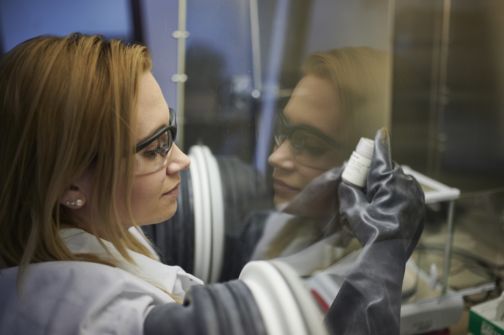 Scientist in safety glasses and gloves holding a small vial inside a lab glove box.