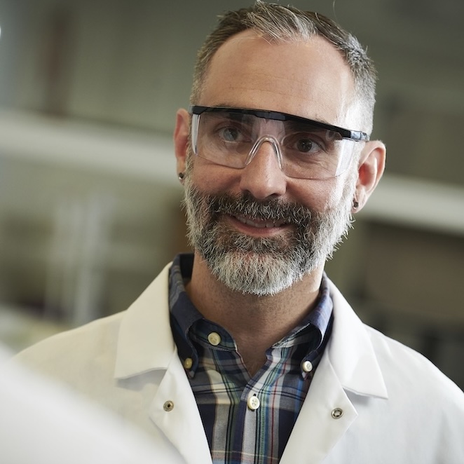 Smiling man with beard and glasses in a white lab coat in a lab setting.