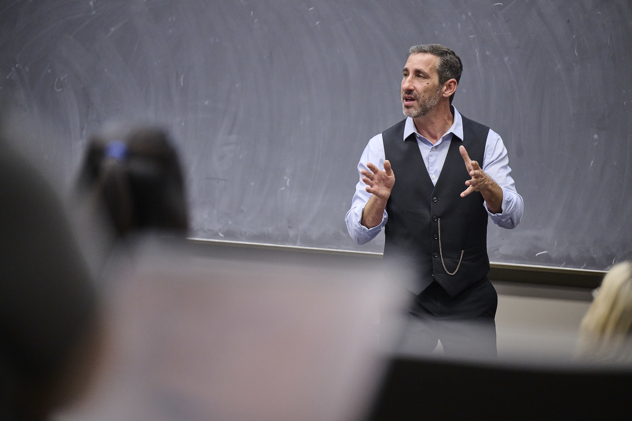 Man in vest and shirt speaking in front of a large chalkboard in a classroom.