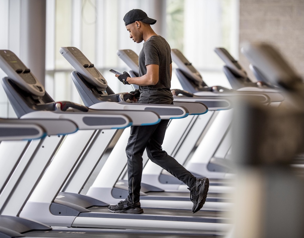 Man in black workout clothes using treadmill while looking at his phone in a gym.