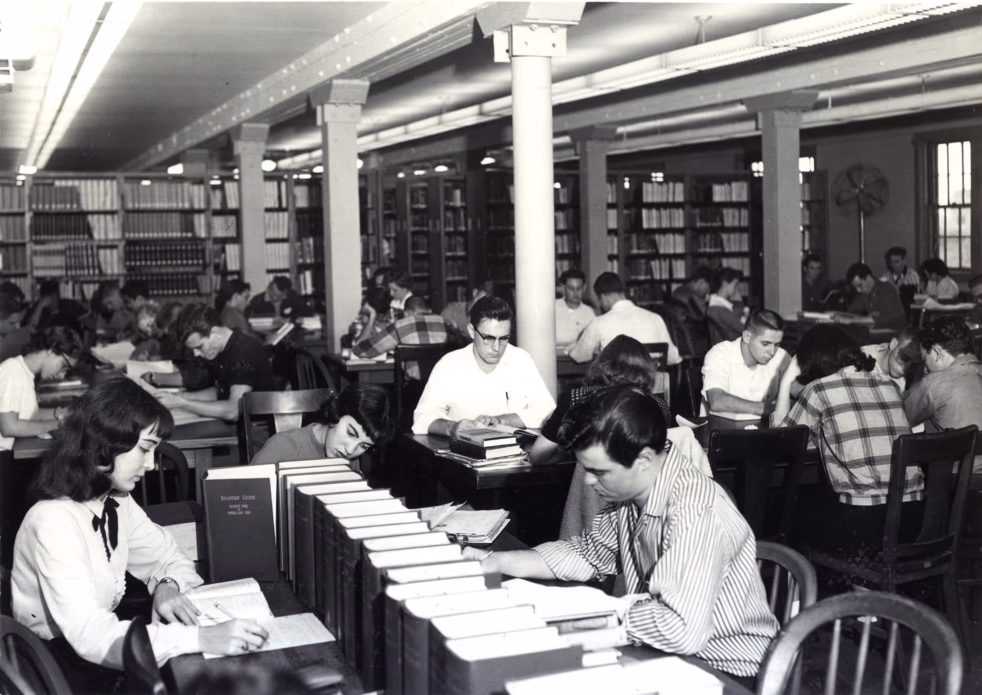 Black and white photo of students studying at tables in a packed library with bookshelves in the background.