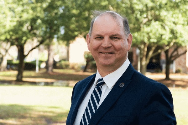 Darrell Kruger. Smiling man in a navy suit and striped tie standing outdoors with trees in the background.