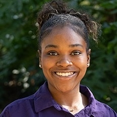 Rozhai Rickmon. Smiling woman with curly hair tied back, wearing a dark purple shirt against a green leafy background.