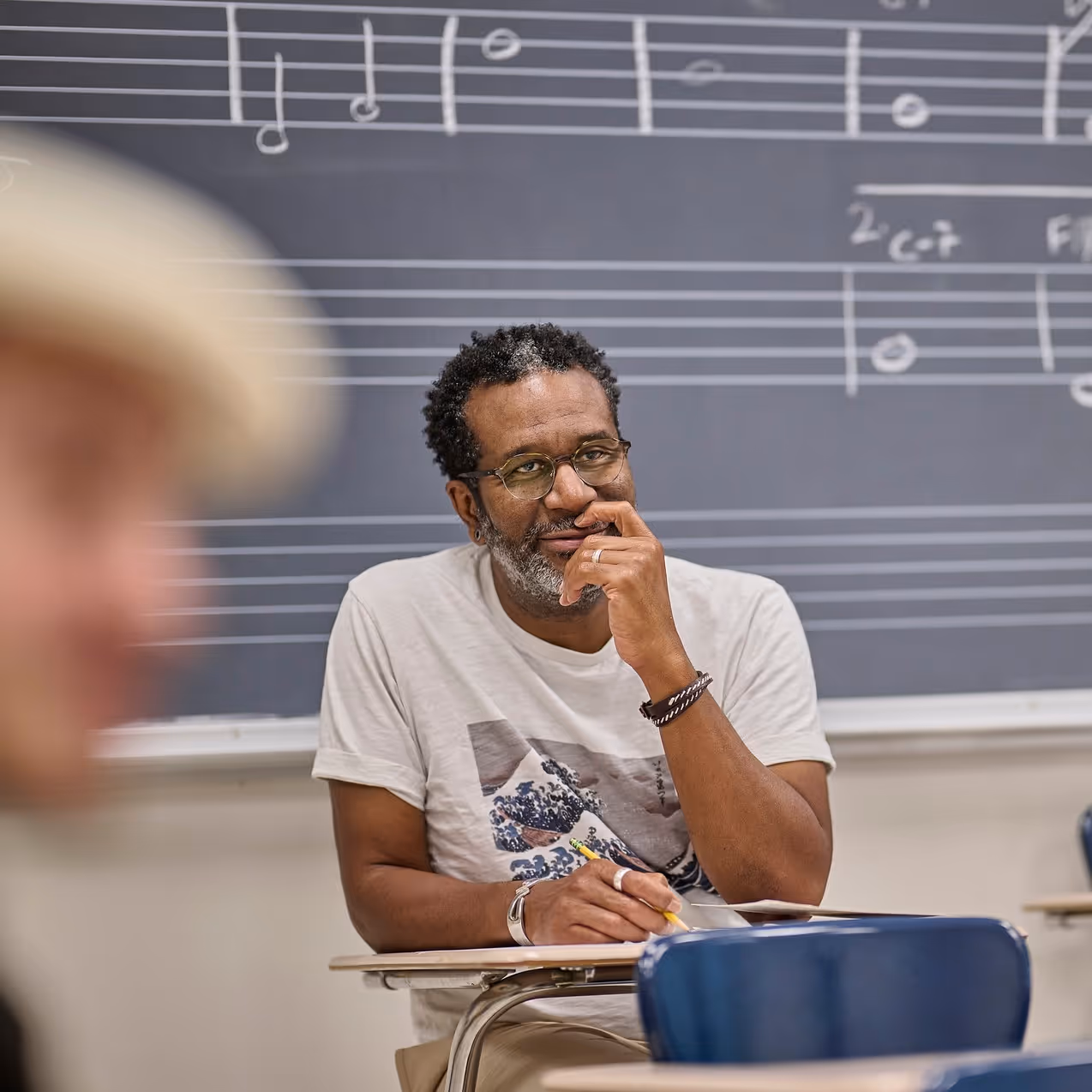 Man with glasses sitting at a desk in front of a chalkboard with music notes, holding a pencil.