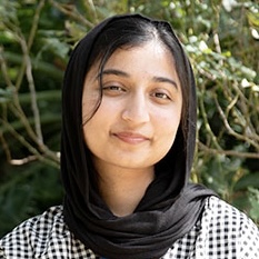 Mahnoor-Cheema. Woman with long dark hair wearing a black headscarf and checkered shirt faces camera outdoors.