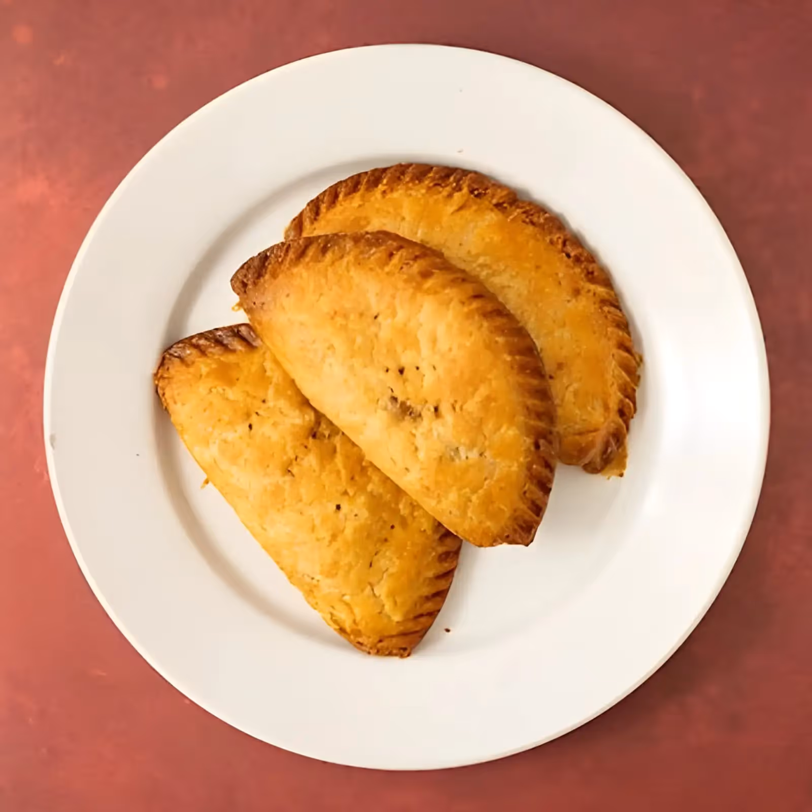 Three golden brown empanadas on a white plate with a reddish background.