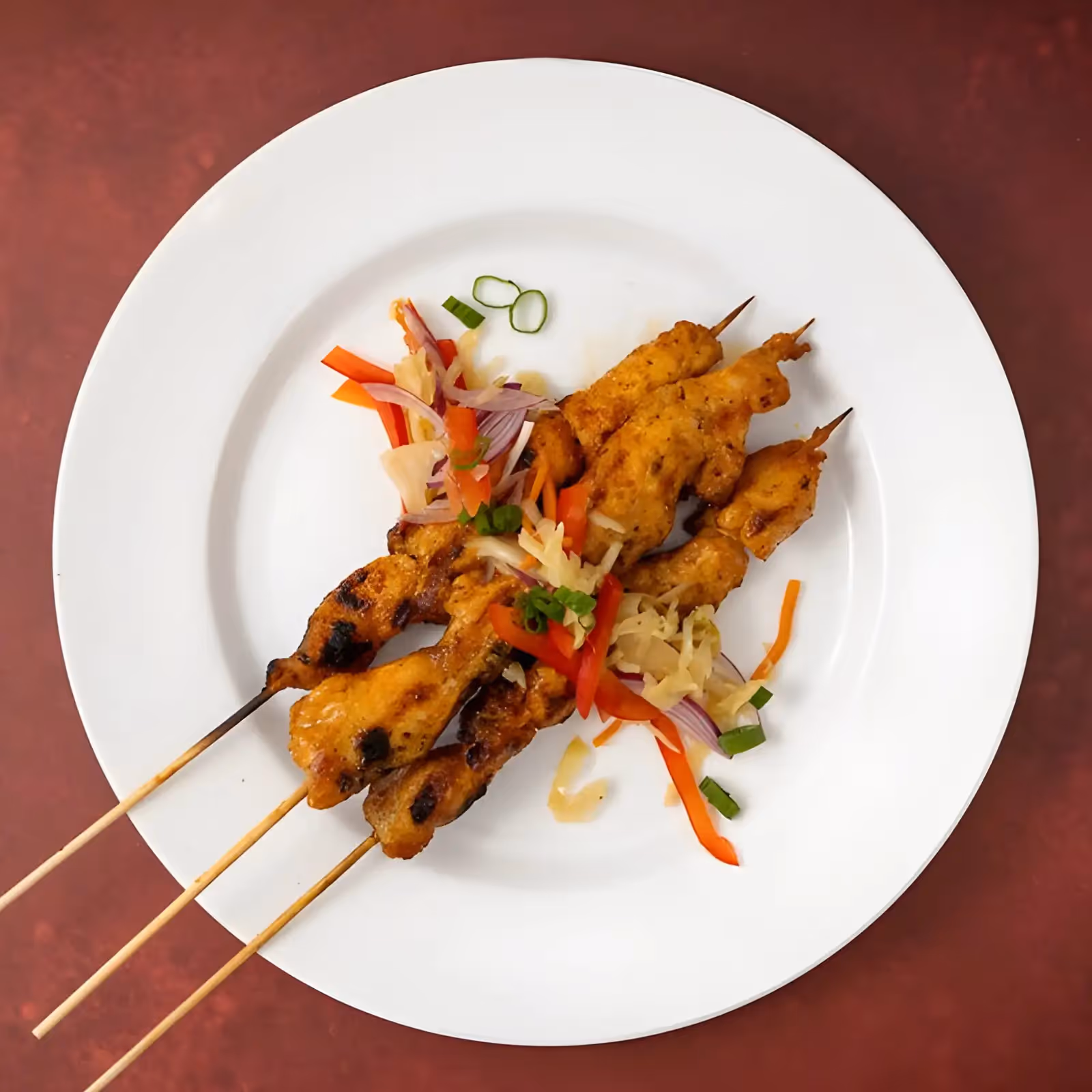Three grilled chicken satay skewers on white plate with sliced vegetables garnish on red table.