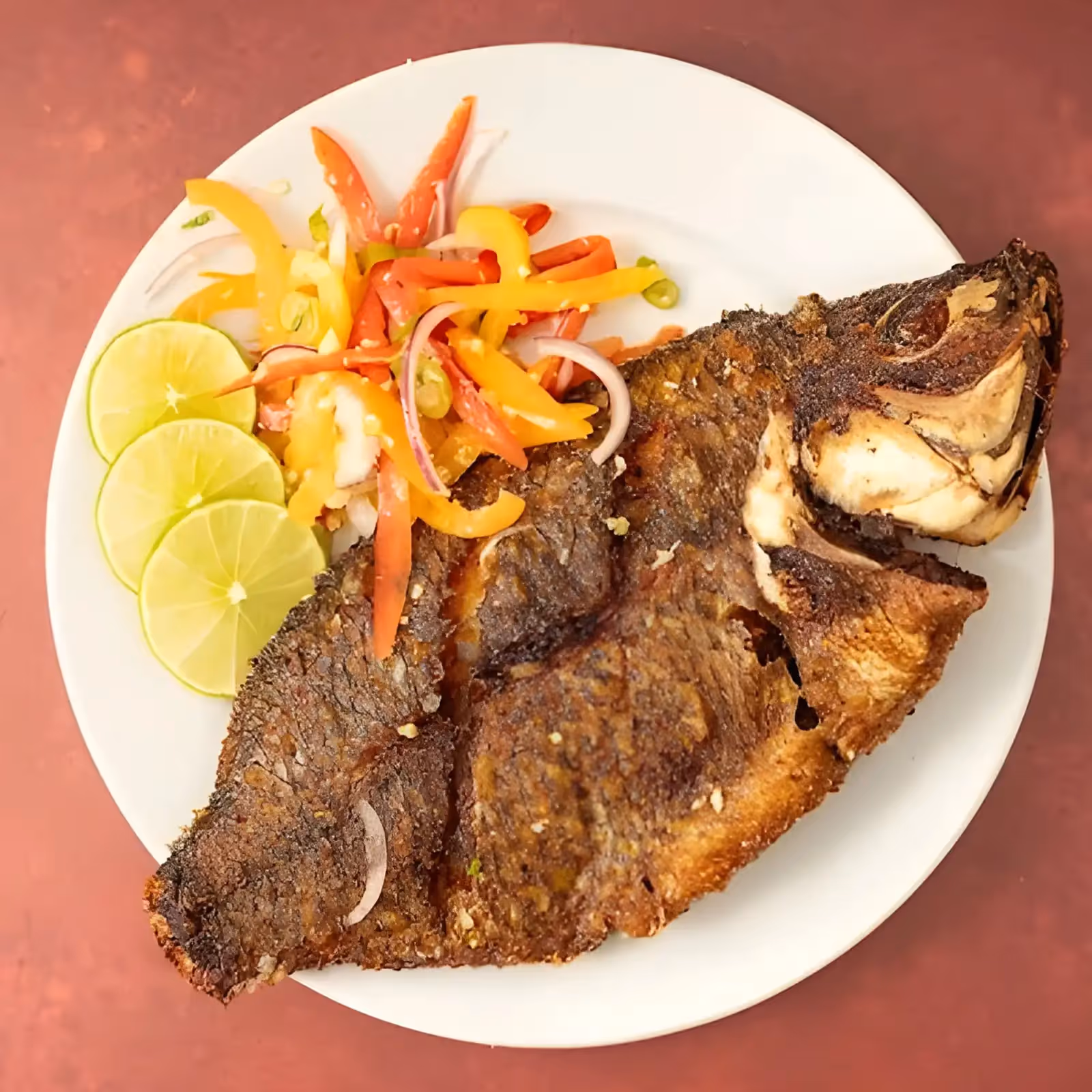 Fried whole fish served on white plate with lime slices and colorful bell pepper salad.
