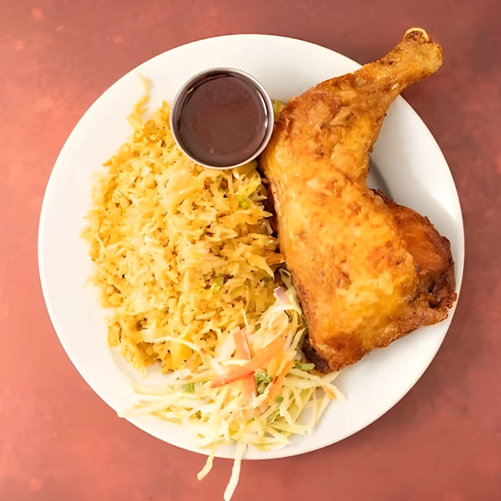 Plate with fried chicken leg, seasoned rice, coleslaw, and a small cup of dark sauce on a red surface.