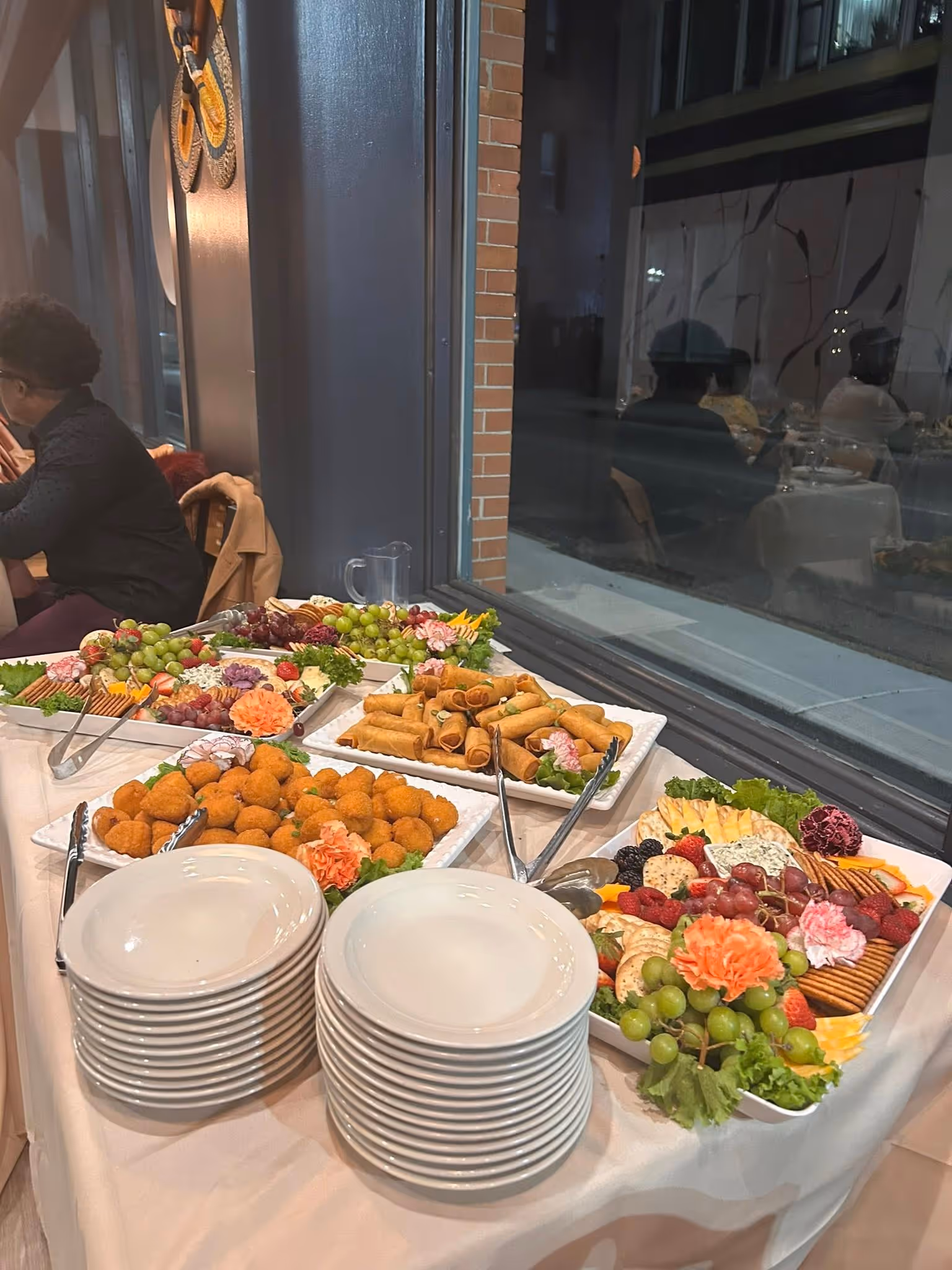 Buffet table with stacks of white plates and trays of spring rolls, fried snacks, and fruit platters.