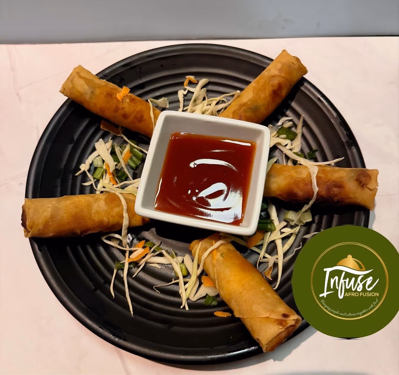 Black plate with five fried spring rolls arranged around a bowl of red dipping sauce and shredded vegetables.