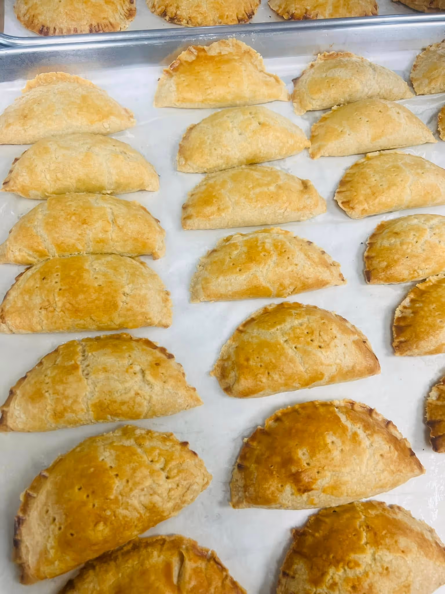 Rows of golden-brown baked empanadas cooling on a white surface and metal tray.