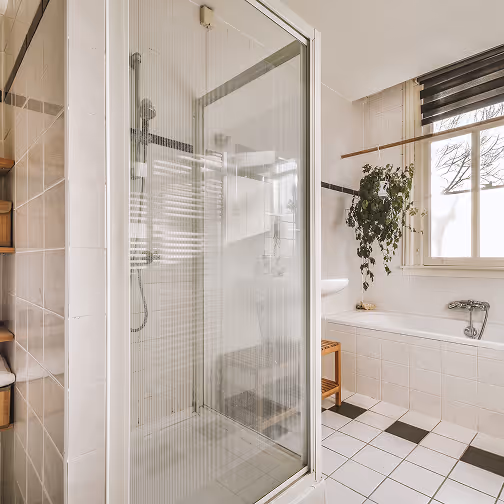 Badkamer met een glazen douchecabine, witte tegels, een ligbad bij het raam met een zittrekje en een hangende groene plant.