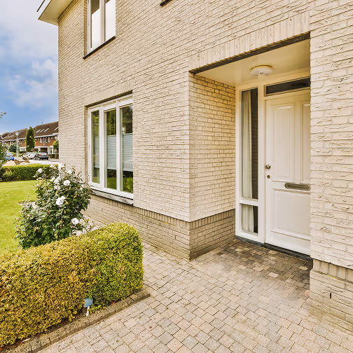Voorkant van een woning met beige bakstenen gevel, witte voordeur en raam, en een nette tuin met groenblijvende struiken en witte bloemen.