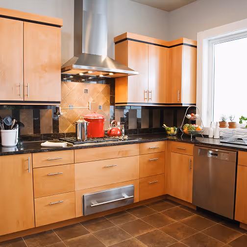 Keuken met licht houten kasten, zwarte werkbladen, roestvrijstalen afzuigkap en apparaten, en een raam rechts.