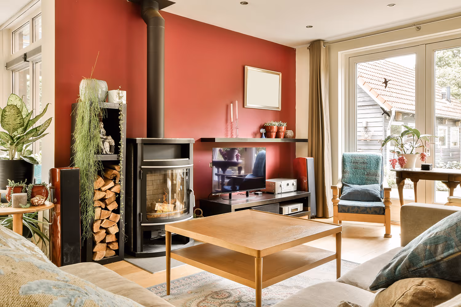 Gezellige woonkamer met houtkachel, stapel brandhout, tv op kast, houten salontafel en grote ramen met uitzicht.