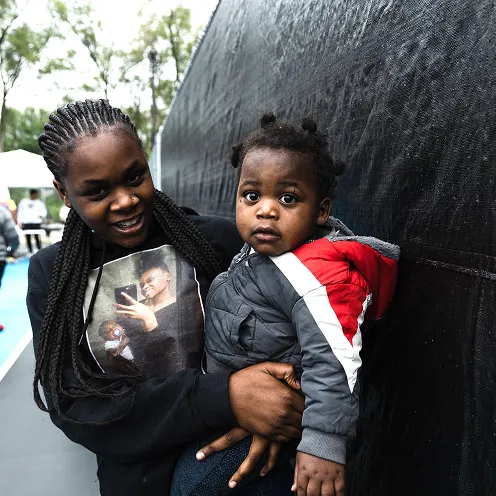 Young woman with braided hair holding a baby boy dressed in a gray and red jacket, both looking at the camera outdoors.