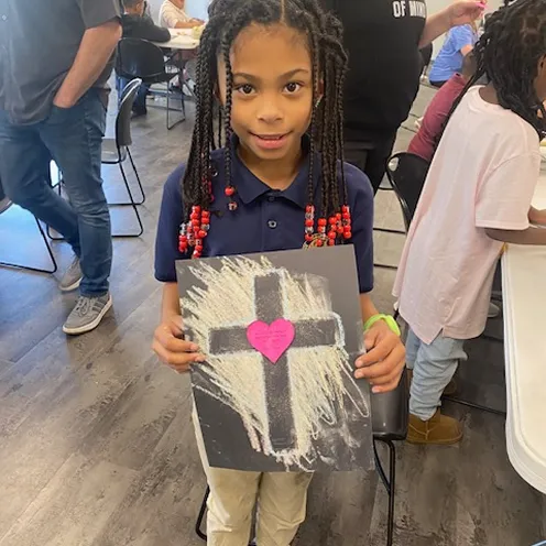 Young girl with braided hair holding a black paper with a chalk-drawn cross and a pink heart in the center.
