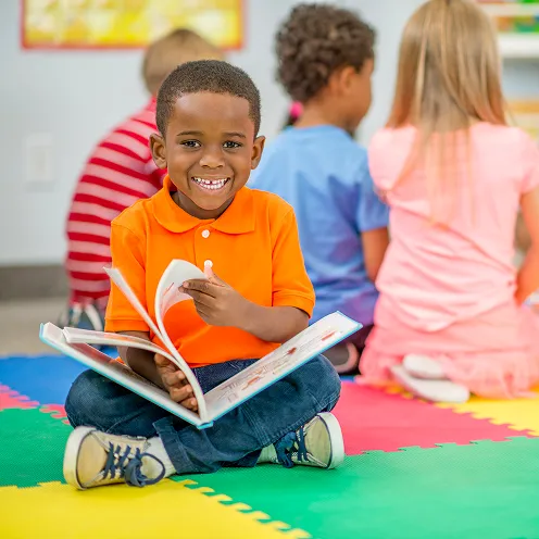 Smiling boy in an orange shirt sitting cross-legged on a colorful foam mat, reading a book in a classroom with other children.