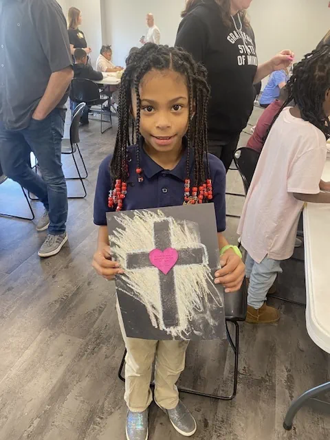 Young girl with braided hair holding a black paper with a chalk-drawn cross and a pink heart in the center.