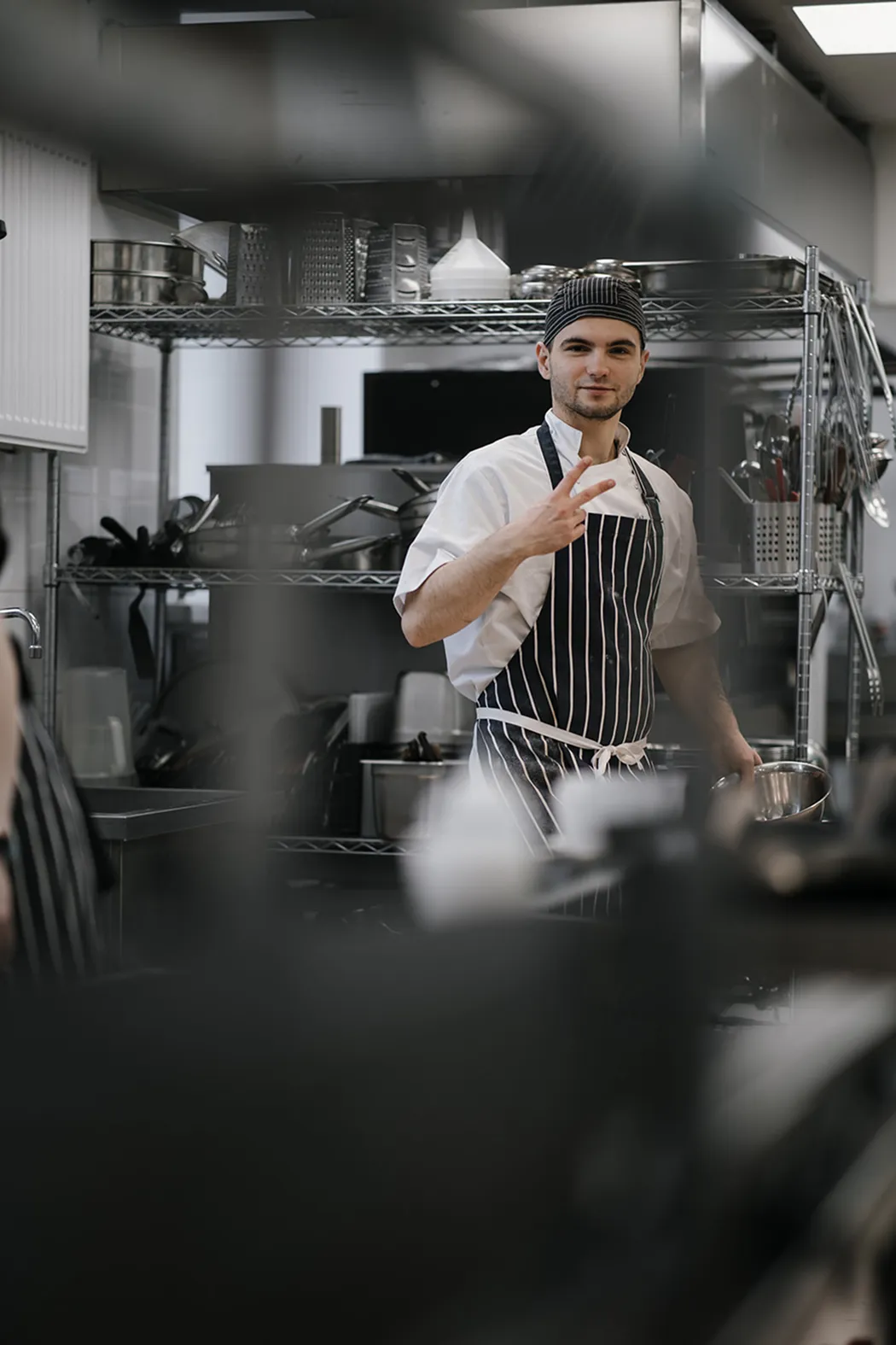 Chef in white uniform and striped apron holding a stainless steel bowl, making a peace sign in a kitchen.