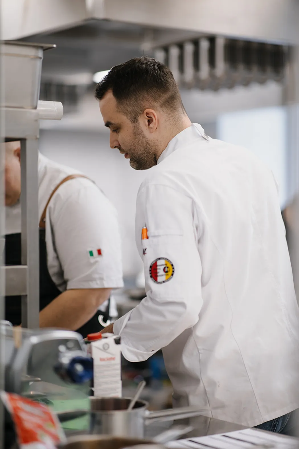 Chef in white uniform preparing food in a professional kitchen with another chef in the background.