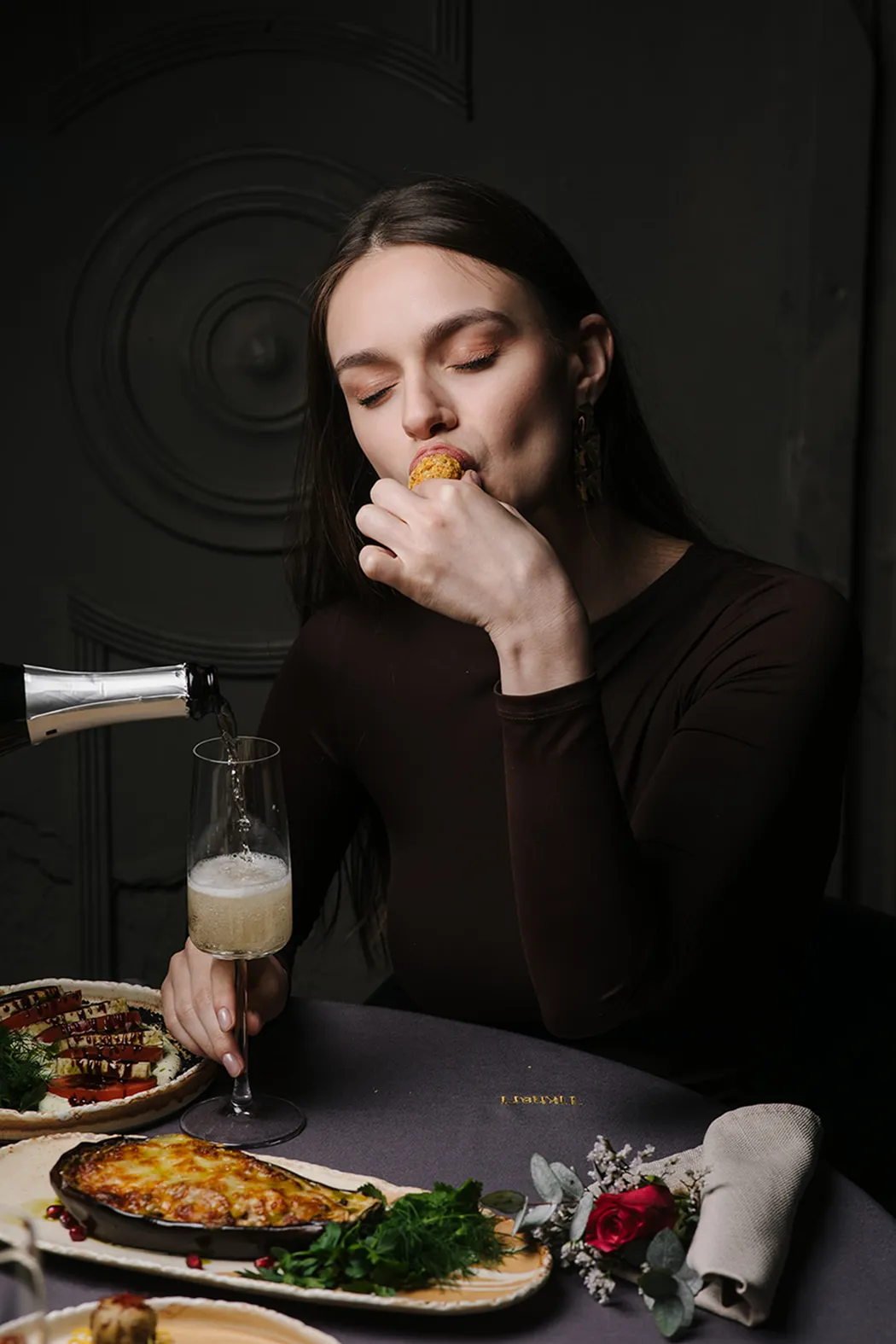 Woman with closed eyes eating a small appetizer while champagne is poured into a flute glass at a table with various dishes and a cloth napkin with a red rose.