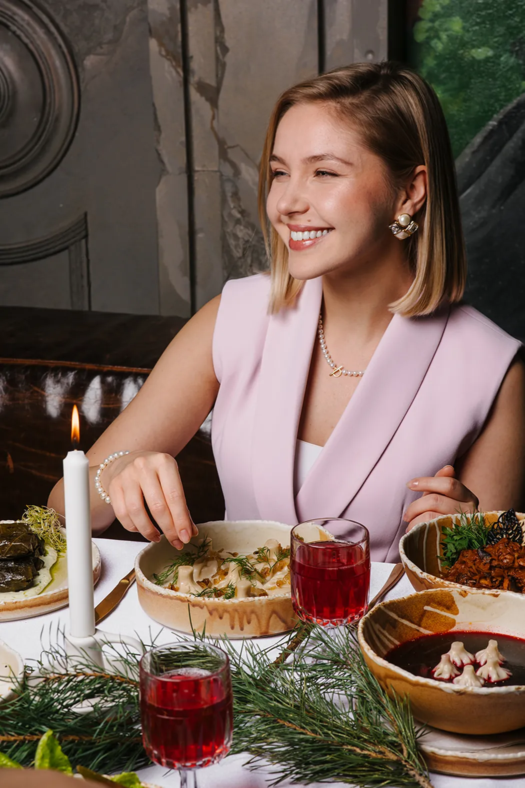 Smiling woman in a light purple sleeveless blazer sitting at a table with festive food, red drinks, and a lit white candle.