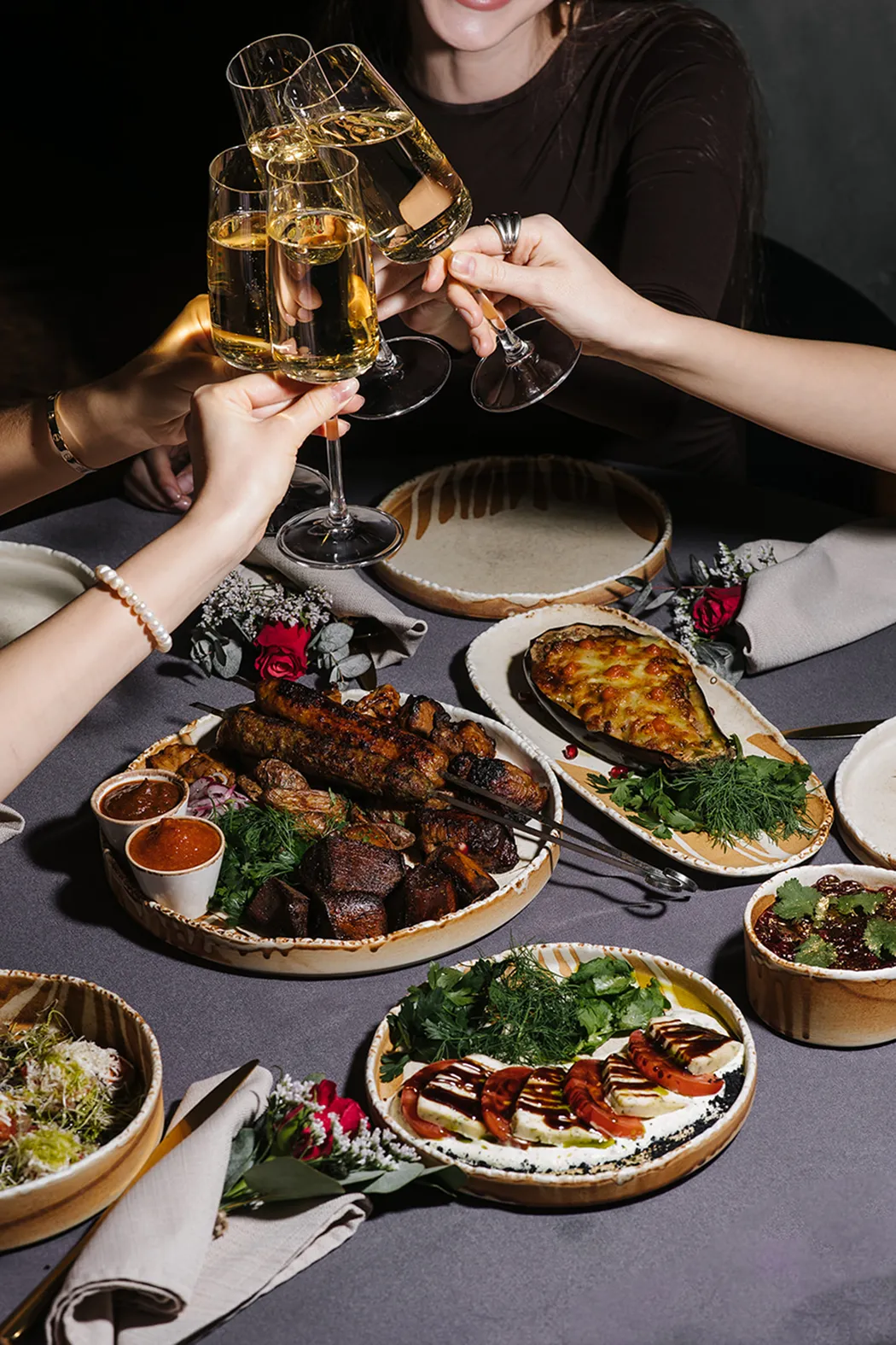 Four hands clinking champagne glasses above a table with plates of grilled meat, stuffed eggplant, tomato and cheese salad, and garnishes.