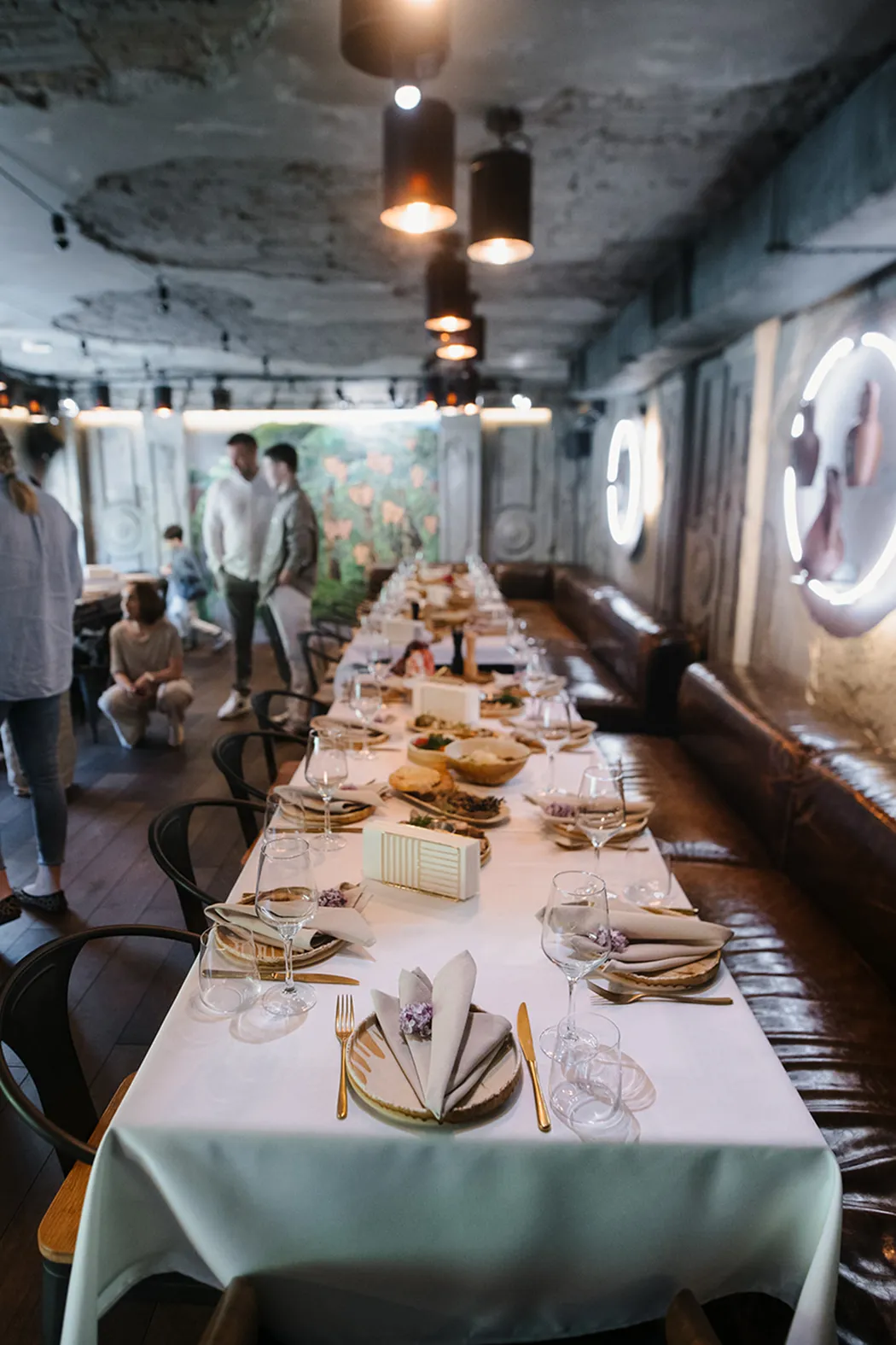 Long dining table with white tablecloth set with plates, folded napkins, gold cutlery, and glassware in a dimly lit rustic restaurant with exposed ceiling and leather bench seating.