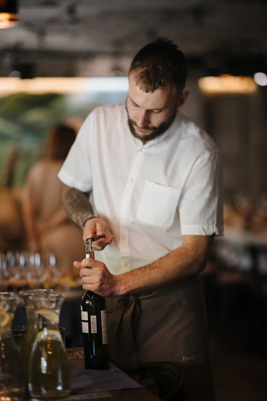 Man in white shirt opening a bottle of wine with a corkscrew in a dimly lit restaurant.