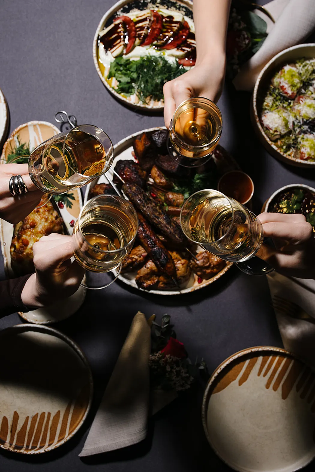 Four hands clinking glasses of white wine over a table with plates of grilled meat, salads, and garnished dishes.
