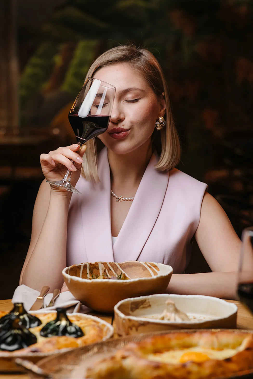 Woman with short blonde hair holding a glass of red wine near her face with eyes closed, seated at a wooden table with dishes of food.