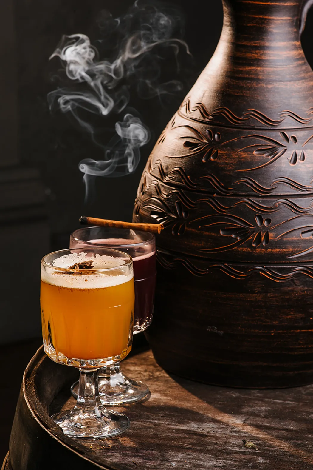 Two glasses of hot spiced beverages with a cinnamon stick and star anise on a wooden surface beside a large decorative clay pot.