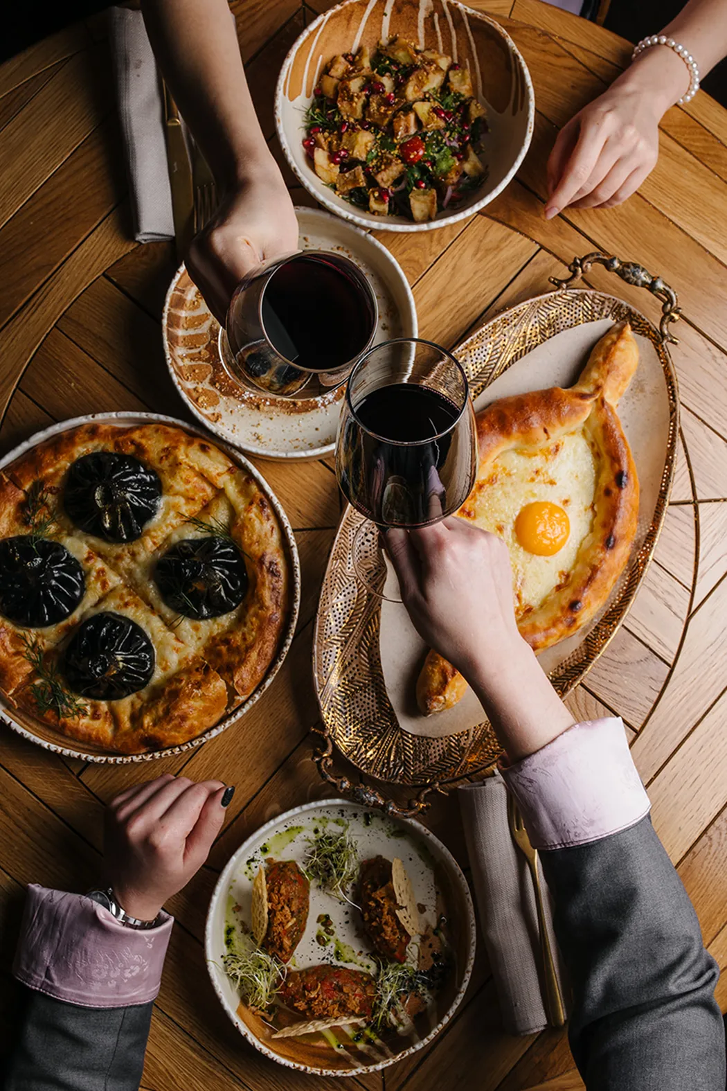 Overhead view of a wooden table with a salad bowl, pizza with black mushrooms, a dish with bread and toppings, a bread boat with an egg yolk, and two people clinking glasses of red wine.