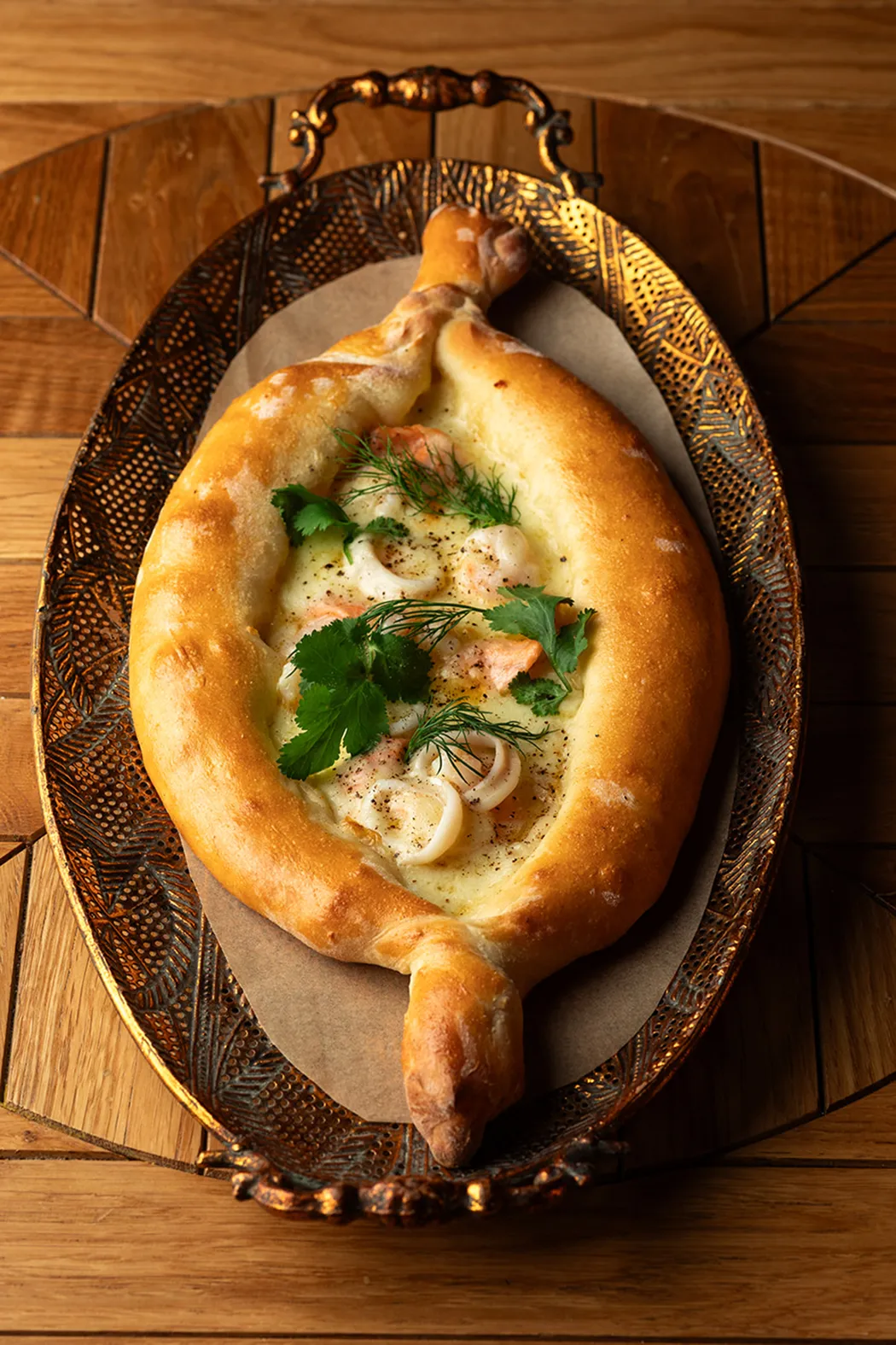 Georgian baked bread boat with cheese, shrimp, calamari, and fresh herbs on a decorative metal tray.
