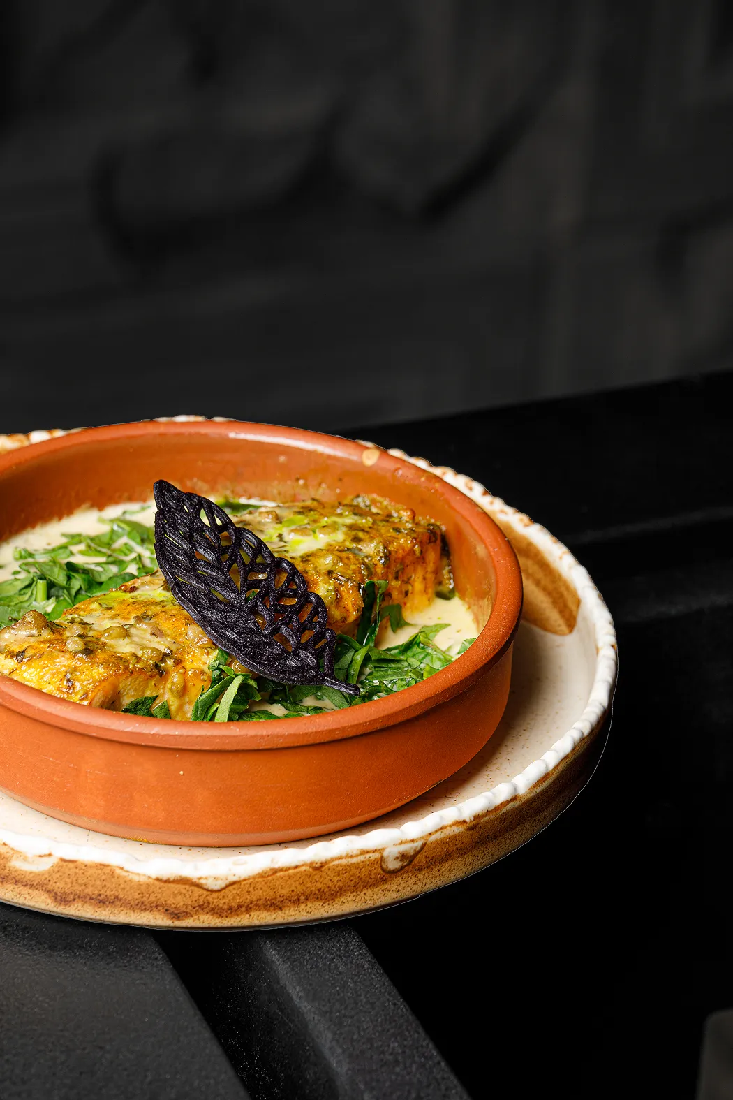 Baked fish fillet in creamy sauce garnished with fresh greens and a black decorative leaf in a brown ceramic dish on a rustic plate.