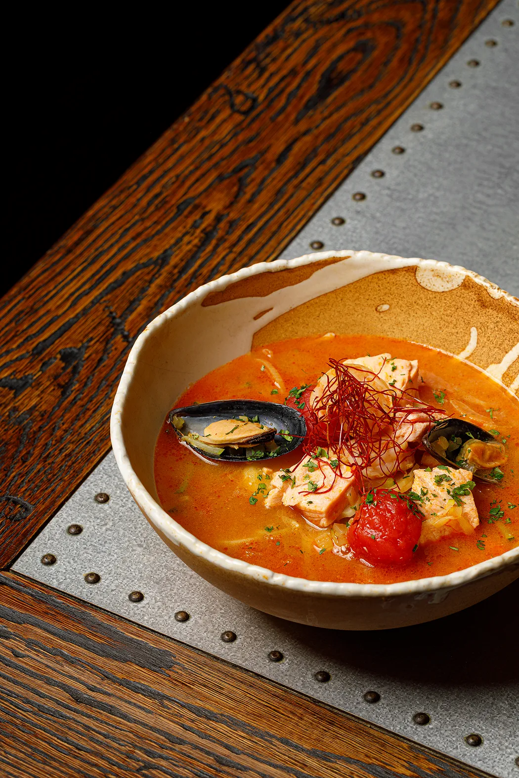 Bowl of seafood stew with mussels, salmon chunks, tomatoes, and garnished with herbs and thin red chili threads.