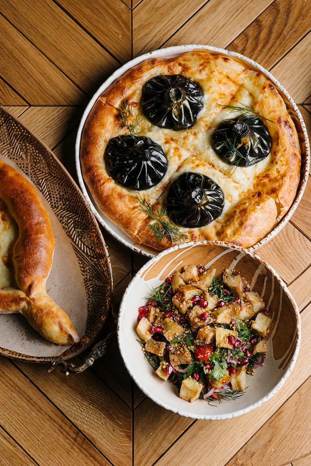 Three dishes on wooden table: a round cheese bread topped with black dumplings, a bowl of mixed salad with pomegranate seeds and vegetables, and a crescent-shaped stuffed bread on a decorative plate.