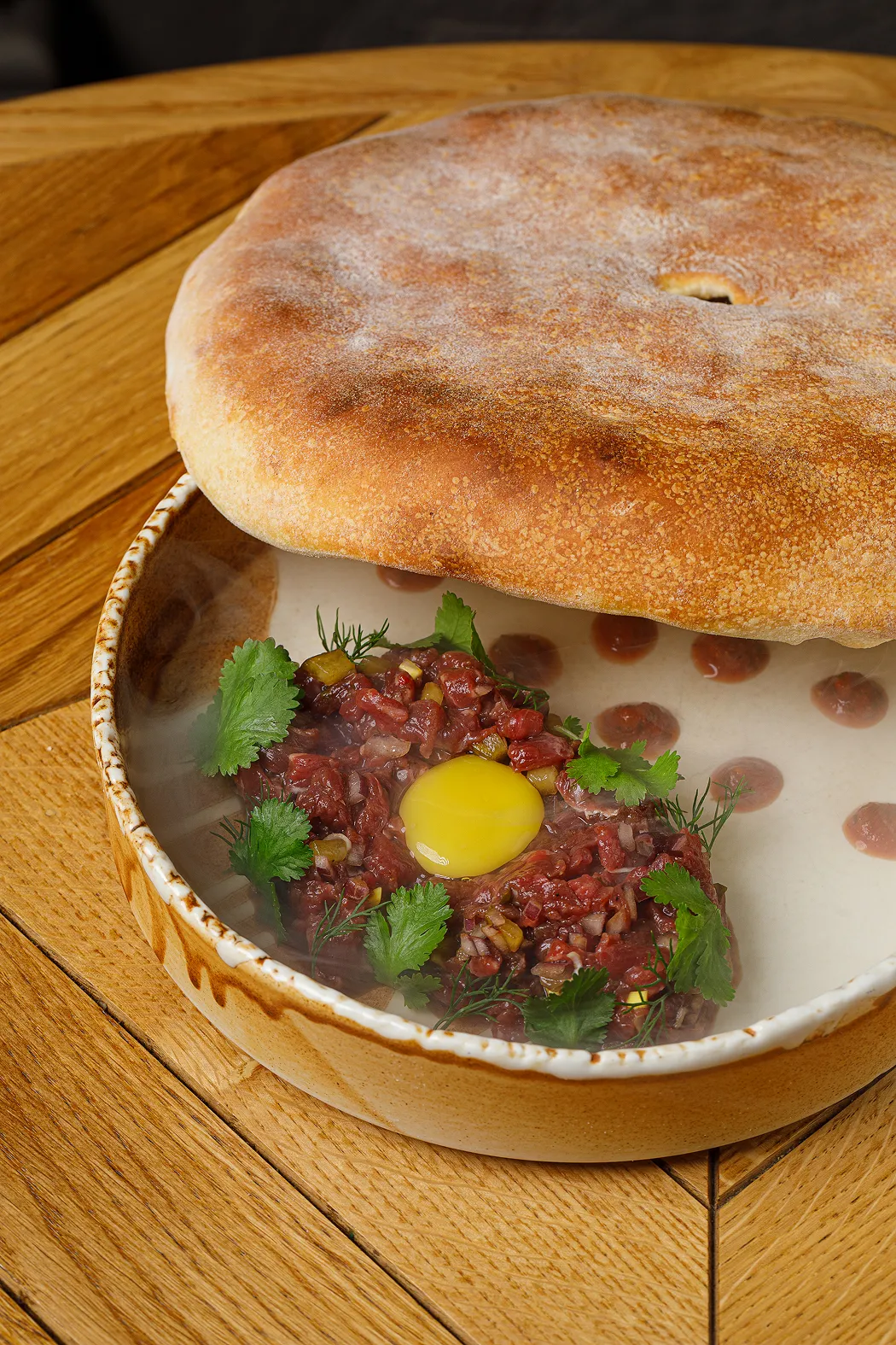 Bowl of steak tartare topped with a raw egg yolk, garnished with fresh herbs, with a toasted bread cover partially lifted.