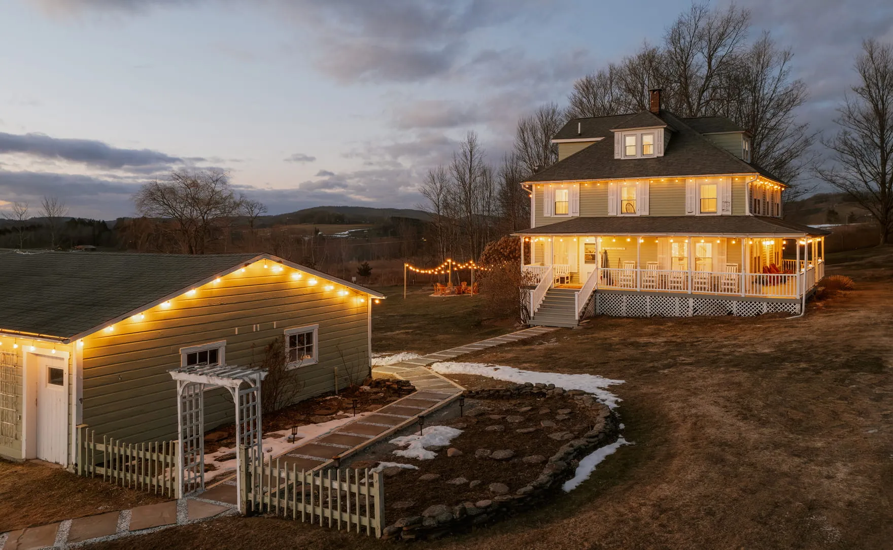 Large two-story house and detached garage decorated with warm string lights at dusk in a rural landscape.